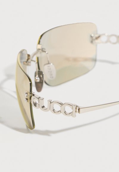Lunettes de soleil sans monture avec des verres marron clair et de fines branches métalliques, avec un logo métallique "GUCCI" sur la branche.
