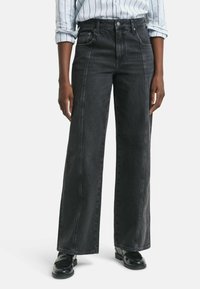Des jeans larges en denim noir présentent une taille haute, des détails de couture verticaux et une texture lisse, associés à des chaussures noires brillantes.