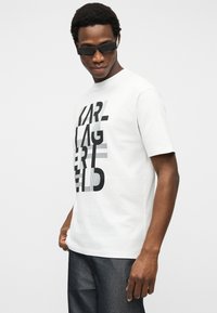 Άνδρας που φοράει μαύρα ορθογώνια γυαλιά ηλίου, λευκό T-shirt με έντονο μαύρο κείμενο και σκούρα τζιν, στέκεται μπροστά σε απλό φόντο.