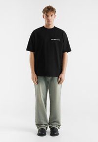 Black cotton t-shirt with short sleeves featuring white text "DON'T WASTE CULTURE" on the chest, paired with light blue baggy jeans and black shoes.