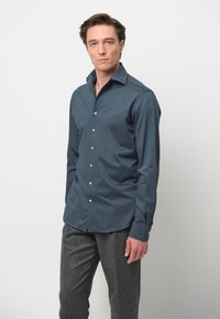 Camicia button-up di un verde scuro con colletto a scorrimento e maniche lunghe. Presenta bottoni bianchi e una superficie liscia. Abbinata a pantaloni grigi.