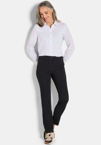 Vrouw met grijs haar, gekleed in een witte blouse, zwarte broek en beige sneakers, staand met handen achter de rug en glimlachend naar beneden.