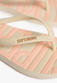 Infradito beige con una fascia testurizzata, con il logo "SUPERDRY" in rilievo. La suola ha uno sfondo rosa con lettere e motivi in rilievo.