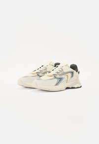 Beige színű sneaker, hálós és velúr textúrákkal. Sárga és zöld kiegészítőkkel rendelkezik, struktúrált talppal és fűzős záródással.