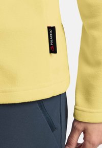 Gelber Fleece-Pullover mit glatter Textur, versehen mit einem sichtbaren schwarzen Polartec-Tag. Er hat einen geraden Saum und ein rundes Bündchen-Design.