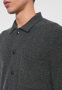 Sötét szürke kötött kardigán, galléros nyakkivágással, négy fekete gombbal és egy mellzsebbelső. A textúrája bordázott.