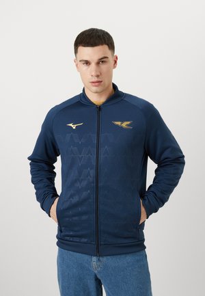 Tume sinine zip-up jakk, mustriga tekstuuriga, kuldsete brändi logo aktsentidega, soonikute mansettidega ja küljetaskutega. Kombineeritud pleekinud siniste teksadega.