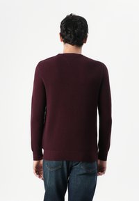 Pull bordeaux avec un motif en tricot texturé, col rond, manches longues et poignets côtelés, associé à un jean bleu. Vue de dos montrée.