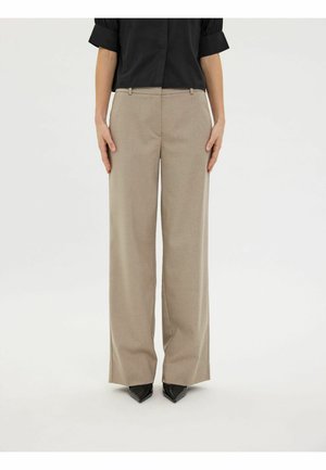 Femme portant un pantalon beige taille haute à jambes larges et des chaussures noires à bouts pointus, debout, les mains détendues le long du corps.
