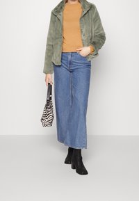 Giacca in pelliccia ecologica verde, dolcevita senape, jeans a gamba larga blu, stivali neri con tacco, tiene in mano una borsa con stampa zebra bianca e nera.