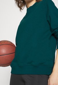 Teal sweatshirt met een ronde hals en ribgebreide zoom, gedragen door een persoon die een basketbal vasthoudt. Fijne stofstructuur, casual pasvorm.