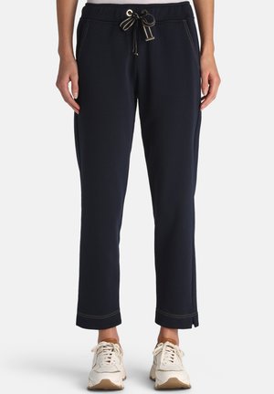 Kvinde iført marineblå bukser med snøre i taljen og sidelommer samt hvide sneakers, stående mod en ensfarvet lys baggrund.