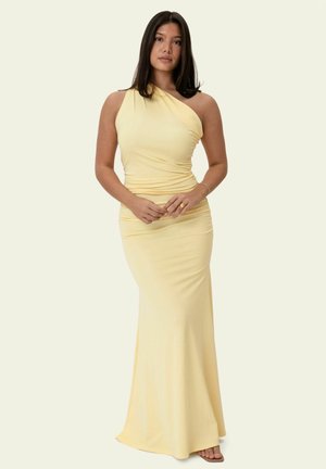 Frau mit glattem dunklem Haar, die ein ärmelloses, blassgelbes, bodenlanges Kleid mit einem Schulterträger trägt, steht vor einem schlichten hellen Hintergrund.