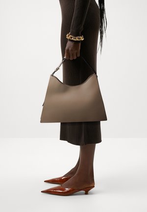 Borsa in pelle marrone con parte superiore curva, lati rigidi e una maniglia corta nera. Indossata con un vestito marrone a coste e tacchi lucidi marroni.