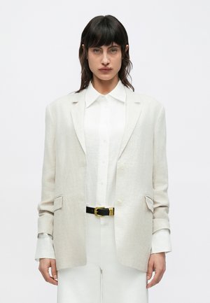 Blazer en lin beige clair avec une coupe décontractée, deux poches avant, deux boutons et une texture subtile, porté sur une chemise blanche boutonnée et une ceinture noire.