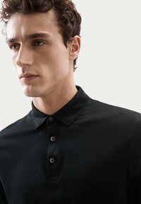 Schwarzes Poloshirt aus glattem Stoff mit einem klassischen Kragen und einer Knopfleiste mit drei Knöpfen. Nahaufnahme des Kragens und des Oberkörpers.