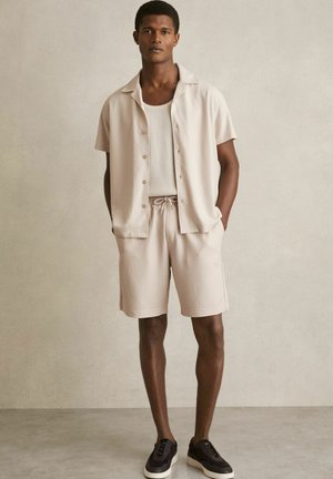 Light beige short-sleeve shirt and matching shorts in ribbed fabric, paired with dark sneakers. Simple design with a button-up front and drawstring waist.