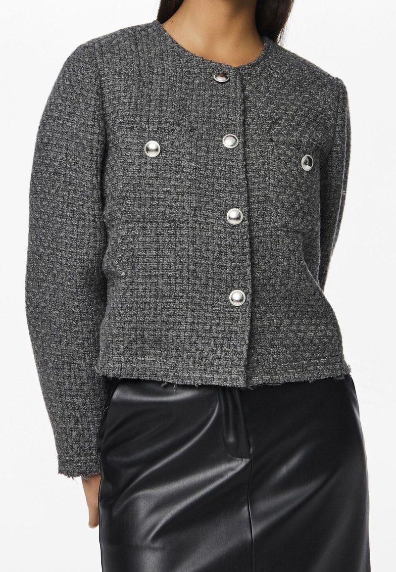 Veste grise texturée boutonnée avec des boutons ronds argentés portée avec une jupe en cuir noire.