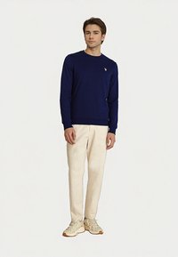 Fiatal férfi sötétkék, hosszú ujjú pólóban, krémszínű nadrágban és bézs sportcipőben áll, sima világos háttér előtt.