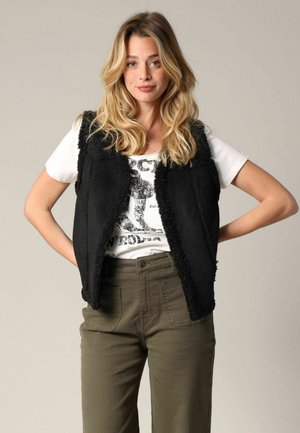 Gilet en fausse fourrure noire avec des franges, porté sur un t-shirt blanc à motif, associé à un pantalon vert olive. Design simple et moderne.