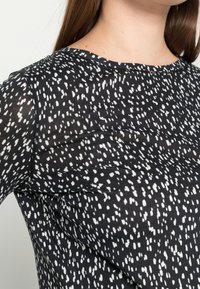 Černý textilní top s náhodným bílým vzorem, kulatým výstřihem a krátkými rukávy, s hladkou strukturou a volným střihem.