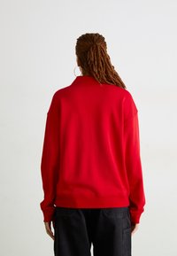 Felpa rossa oversize con colletto alto, polsini e orlo a coste. Il materiale sembra morbido con una texture liscia. Sotto sono indossati pantaloni neri.