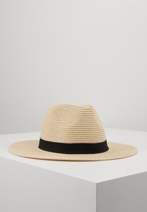 Beżowy szerokoklapowy kapelusz fedora z czarną opaską, prezentowany na białej powierzchni na jednolitym jasnoszarym tle.