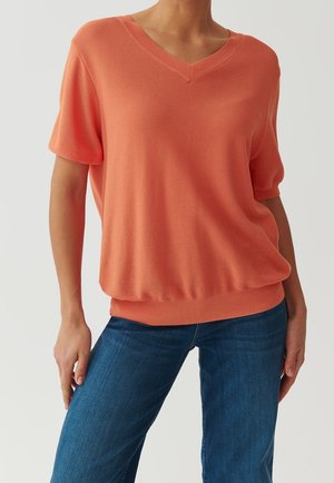 Kobieta ubrana w koralowy krótki sweter z dekoltem w serek, w zestawieniu z niebieskimi dżinsami, stoi z jedną ręką na biodrze na jednolitym tle.