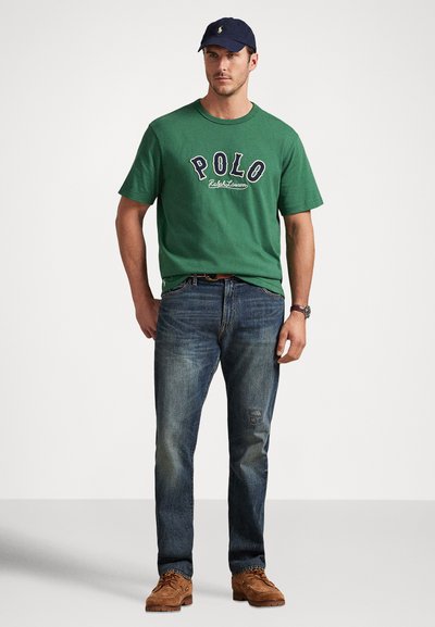 Vyras, vilkintis žalią Polo Ralph Lauren marškinėlius, mėlynas džinsus, rudus batus, juodą laikrodį ir tamsiai mėlyną kepurę, stovi priešais baltą foną.