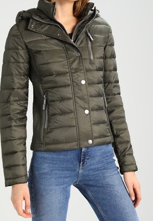 Frau trägt eine taillierte, olivgrüne Steppjacke mit Reißverschluss und Druckknöpfen, kombiniert mit blauen Jeans, vor einem einfarbigen Hintergrund.