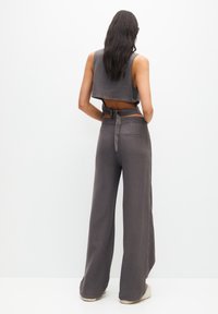 Mujer con cabello largo y oscuro, vistiendo un top corto sin mangas gris y pantalones de pierna ancha gris de cintura alta, de pie frente a un fondo blanco.