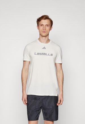 T-shirt de sport blanc avec le graphique "LesMILLS" en gris, à manches courtes, associé à un short noir à motifs avec des lignes courbes.