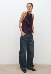Deep burgundy halter top with draped neckline paired with wide-leg dark denim jeans. Model stands with hands in pockets, wearing black boots.
