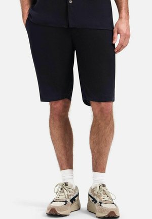 Man draagt zwarte shorts, witte sokken en beige sneakers en staat tegen een effen lichte achtergrond.