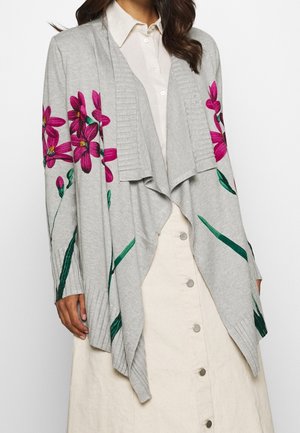 Frau trägt eine graue Strickjacke mit pinker Blumenstickerei über einem weißen Hemd und einem cremefarbenen Rock mit metallischen Knöpfen.