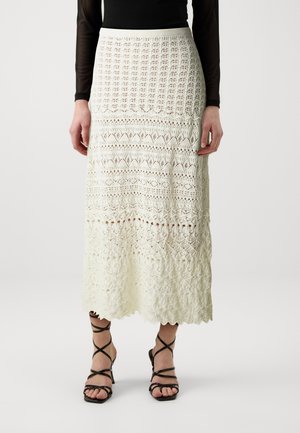 Femme portant une longue jupe blanche en crochet avec des motifs et des talons noirs à brides, debout devant un fond uni.