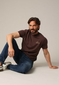 T-shirt marron à manches courtes avec un petit logo doré, associé à un jean bleu foncé et des chaussures de sport blanches. Modèle assis contre un fond neutre.