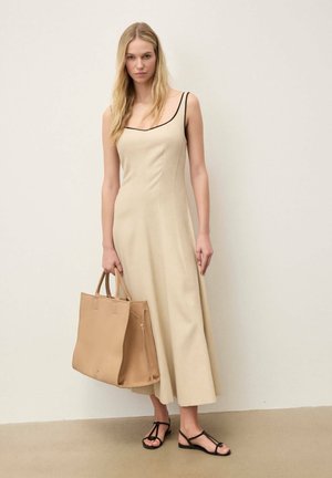 Ξανθιά γυναίκα με αμάνικο μπεζ φόρεμα με μαύρη επένδυση, κρατώντας τσάντα ώμου καφέ ανοιχτού χρώματος, φοράει μαύρα σανδάλια με λουράκια, στέκεται μπροστά σε ανοιχτόχρωμο τοίχο.