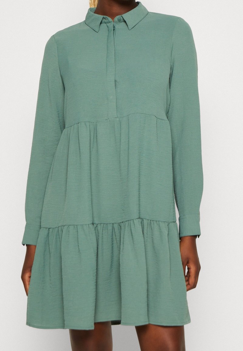Robe longue à manches longues vert sauge à étages avec col et boutons dissimulés à l'avant portée par une personne à la peau foncée.