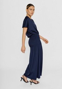 Navyblauwe jurk met een losse pasvorm, korte mouwen en een smocked taille, gecombineerd met zwarte sandaaltjes met bandjes. Zacht, lichtgewicht stof, vloeiende ontwerp.