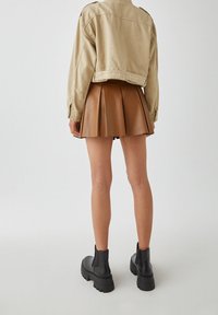 Skórzana, brązowa spódnica plisowana z wysokim stanem, zestawiona z krótką beżową kurtką i czarnymi, masywnymi botkami za kostkę. Gładka tekstura i strumieniowy design.