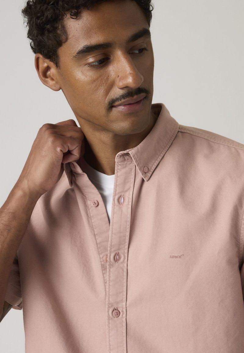 Mann mit Schnurrbart richtet den Kragen eines hellrosa Hemdes über einem weißen T-Shirt, schaut leicht nach unten und zur Seite.