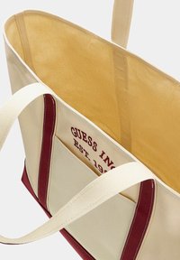 Canvas schoudertas in crèmewit met bordeaux accenten; voorzien van stevige handvatten, een geprinte logo in rood en een ruime, effen binnenkant.