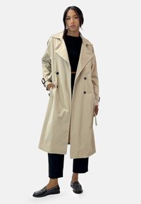 Ljusbeige trench coat med stora slag, dubbelknapprad och sidofickor. Matchas med svart kort topp och byxor.
