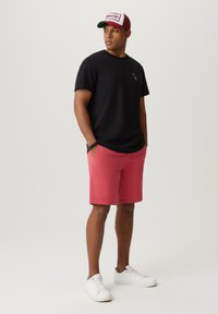 Czarna koszulka, różowe szorty, białe sneakersy. Koszulka ma małe logo; czapka jest czerwona, biała i zielona z napisem z przodu.