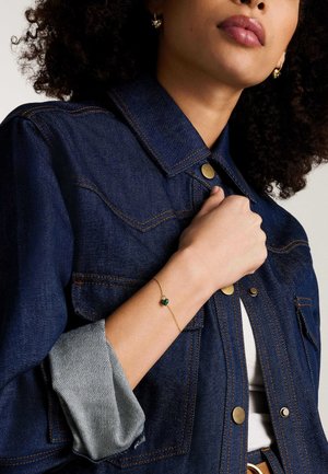 Donna che indossa una giacca di jeans blu scuro con maniche arrotolate e gioielli dorati, tra cui orecchini a forma di cuore e un braccialetto con un ciondolo a cuore verde.