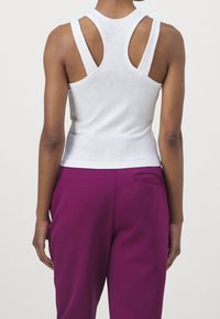 Personne portant un haut blanc sans manches en maille côtelée avec des détails découpés dans le dos et un pantalon violet, debout devant un fond blanc uni.