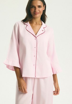 Woman wearing light pink pajama set with red trim, button-up shirt with 3/4 ruffled sleeves, and small cherry embroidery on collar.