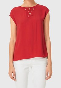 Rød kortærmet bluse med rund hals og indviklet udskæring ved halsen; glat stof og afslappet pasform.