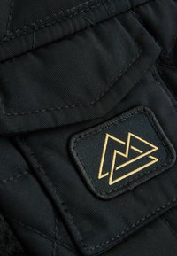 Czarna pikowana tkanina z teksturową powierzchnią. Zawiera złotą naszywkę z logo o geometrycznym wzorze, umieszczoną na klapie.